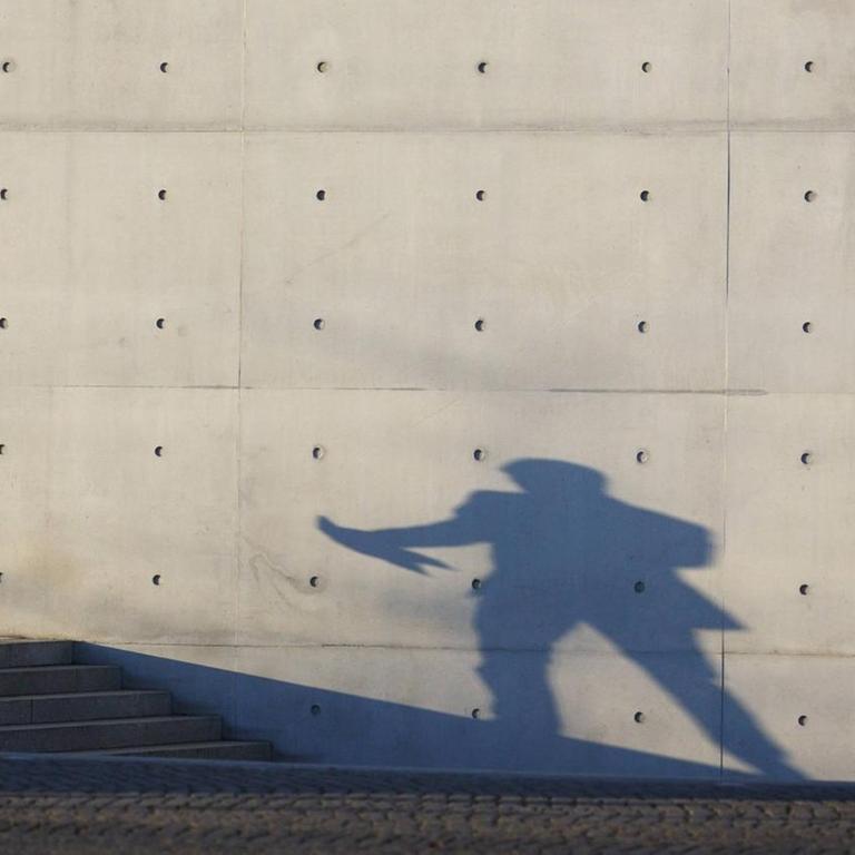 Eine Mann im Anzug telefoniert vor einer Betonwand und wirft dabei einen Schatten. 