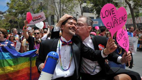 Zwei Männer küssen sich zwischen einer Regenbogen-Flagge und einem herzförmigen Plakat.