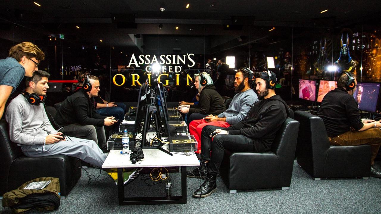 Journalisten sitzen mit Controllern und Kopfhörern auf niedrigen Sesseln um einen Tisch mit Bildschirmen herum. im Hintergrund ist groß der Schriftzug "Assassin's Creed Origins" zu lesen.