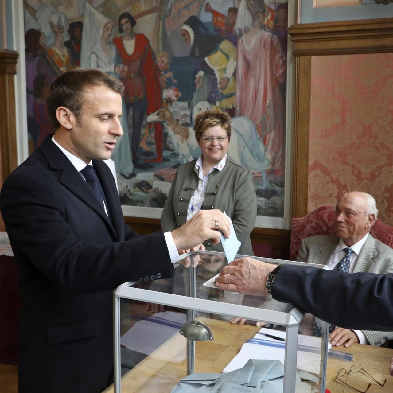Emmanuel Macron steckt den verschlossenen Umschlag mit seinem Wahlzettel in eine transparente Wahlurne. Auf der anderen Seite kontrolliert ein Wahlhelfer ven Vorgang. 