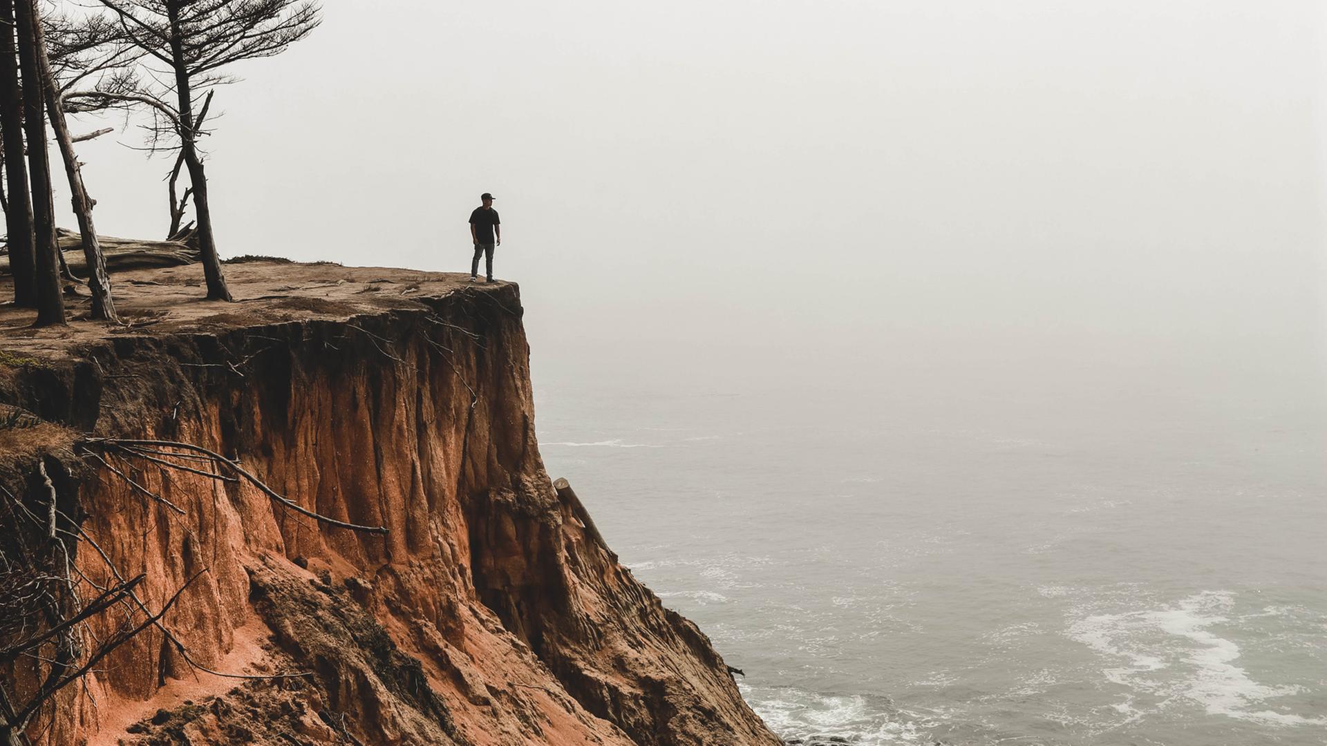 Ein Mensch steht an einer Klippe und schaut aufs nebelige, graue Meer hinaus. Ein Mensch steht an einer Klippe und schaut aufs nebelige, graue Meer hinaus.