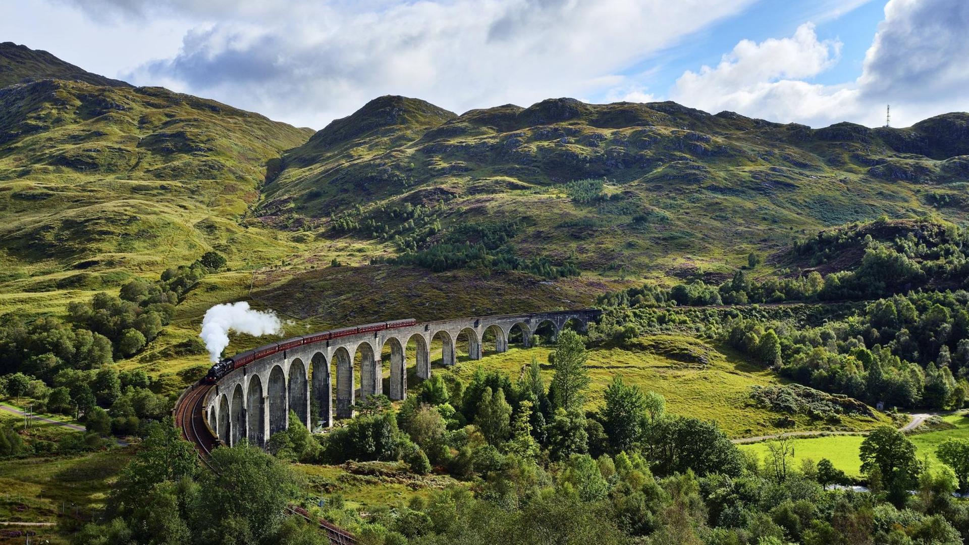 Eine Dampflock fährt über das Glenfinnan Viadukt vor grüner Highlands Landschaft und wolkig blauem Himmel. Eine Dampflock fährt über das Glenfinnan Viadukt vor grüner Highlands Landschaft und wolkig blauem Himmel.
