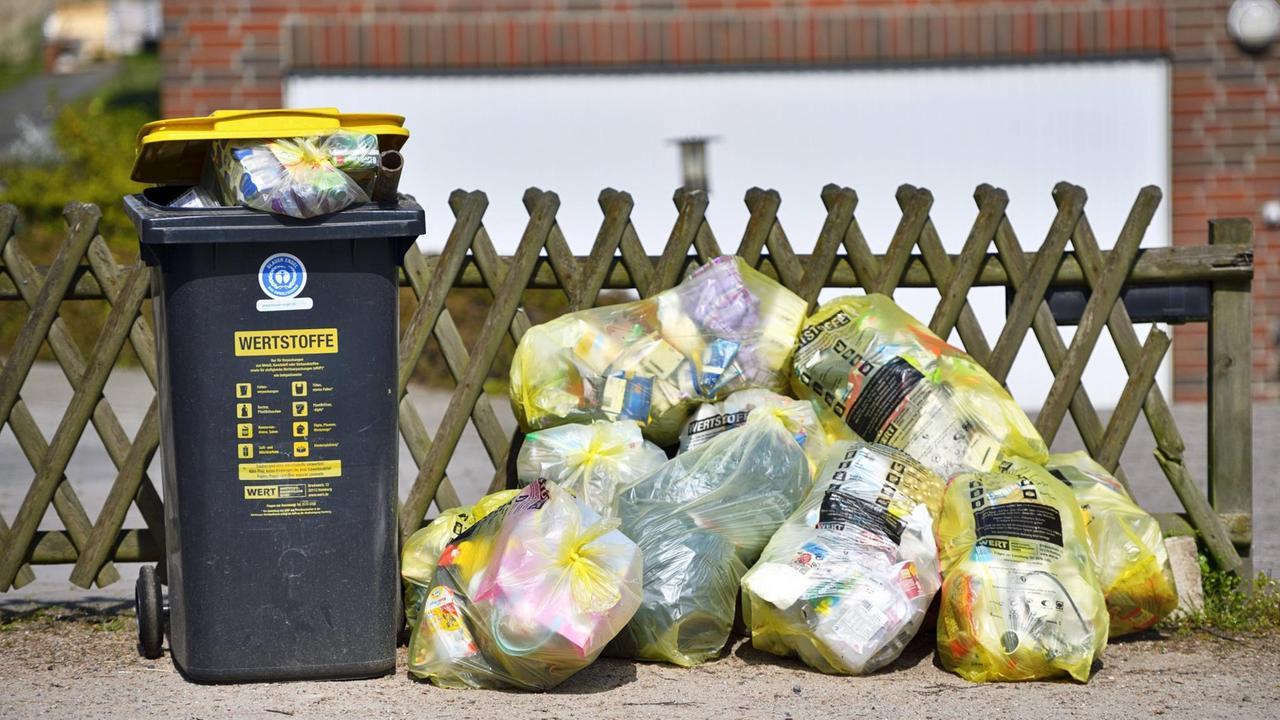 Wertstoffabholung in Hamburg: eine Wertstofftonne vor einem Gartenzaun, daneben ein Stape Gelber Säcke Wertstoffabholung in Hamburg: eine Wertstofftonne vor einem Gartenzaun, daneben ein Stape Gelber Säcke