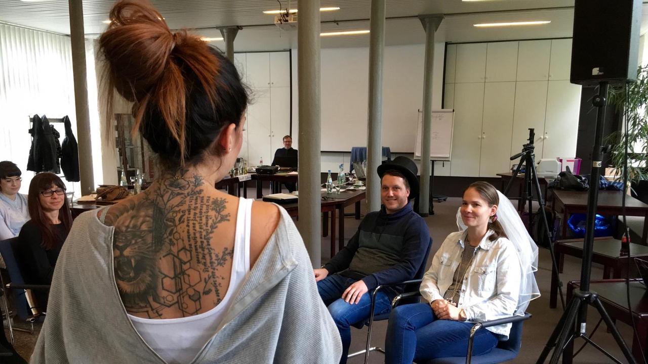 Ein als Brautpaar verkleidetes Paar sitzt vor einer Rednerin. Ein als Brautpaar verkleidetes Paar sitzt vor einer Rednerin.