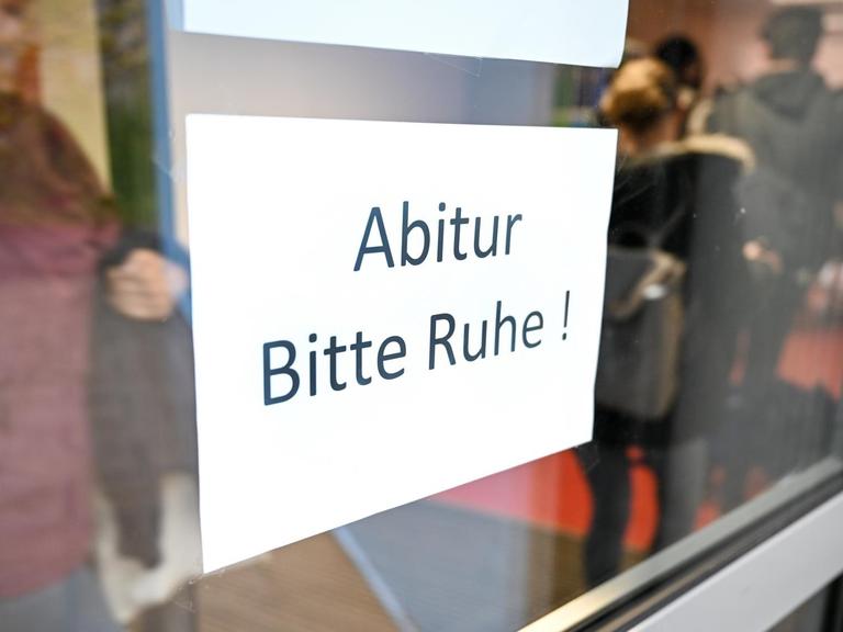 Abiturienten gehen in eime Gymnasium kurz vor Beginn des Abiturs im Fach Deutsch zu ihrem Prüfungsraum an einem Schild vorbei, auf dem "Abitur Bitte Ruhe!" steht.