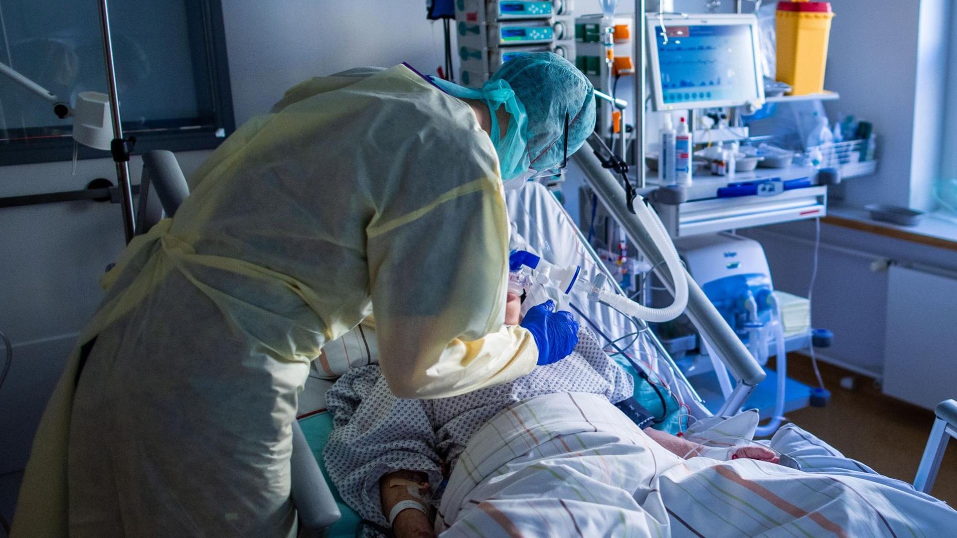 Ein Krankenhausmitarbeiter in Schutzkleidung steht am Bett eines Intensivpatienten. Ein Krankenhausmitarbeiter in Schutzkleidung steht am Bett eines Intensivpatienten.