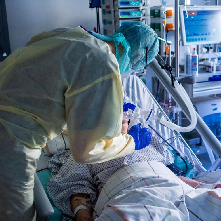 Ein Krankenhausmitarbeiter in Schutzkleidung steht am Bett eines Intensivpatienten.