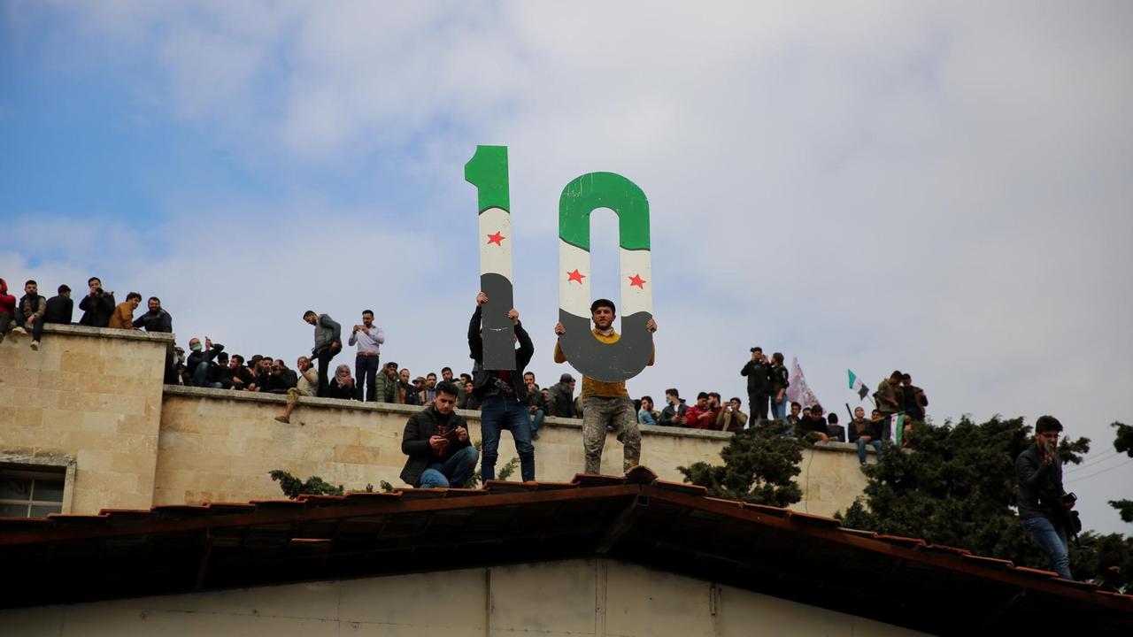Mehrere Menschen haben sich auf Dächern versammelt, zwei halten die zahlen 1 und 0 in den Flaggenfarben der Opposition (grün, weiß, schwarz, drei rote Sterne in der Mitte) hoch.
