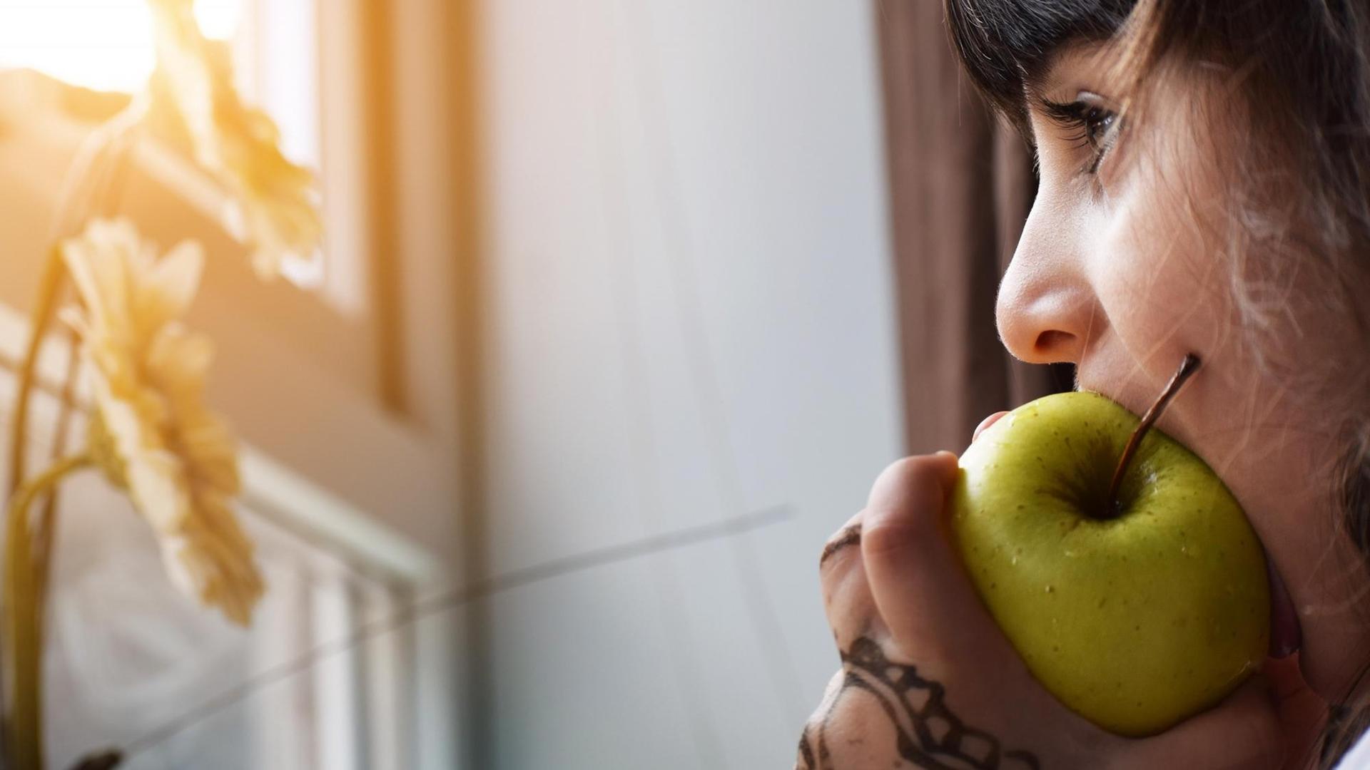 Eine junge Frau isst einen Apfel während sie am Fenster steht. (Symbolbild) Eine junge Frau isst einen Apfel während sie am Fenster steht. (Symbolbild)