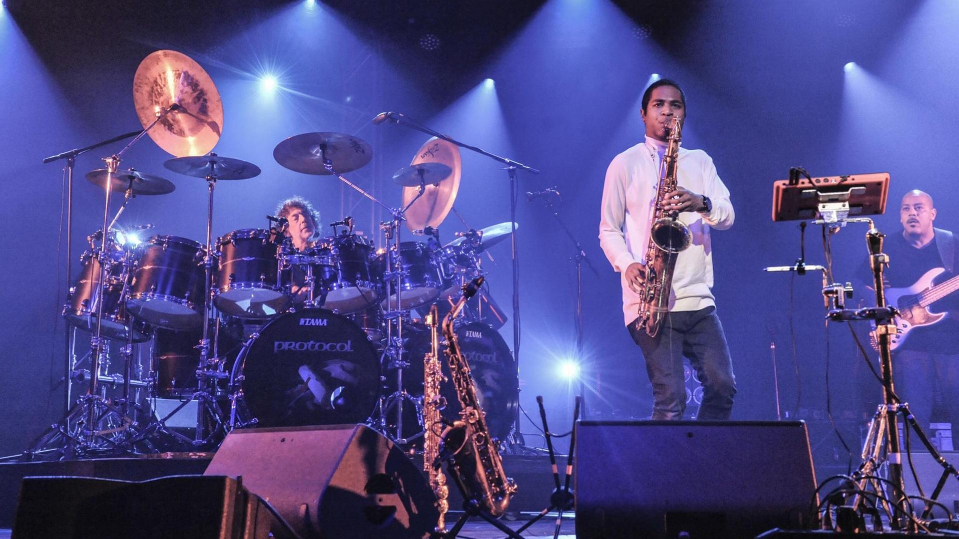 Ein Schlagzeuger (hinten links, ein Saxfonist (Mitte) und ein Bassist (rechts) stehen auf einer Bühne im Scheinwerferlicht.