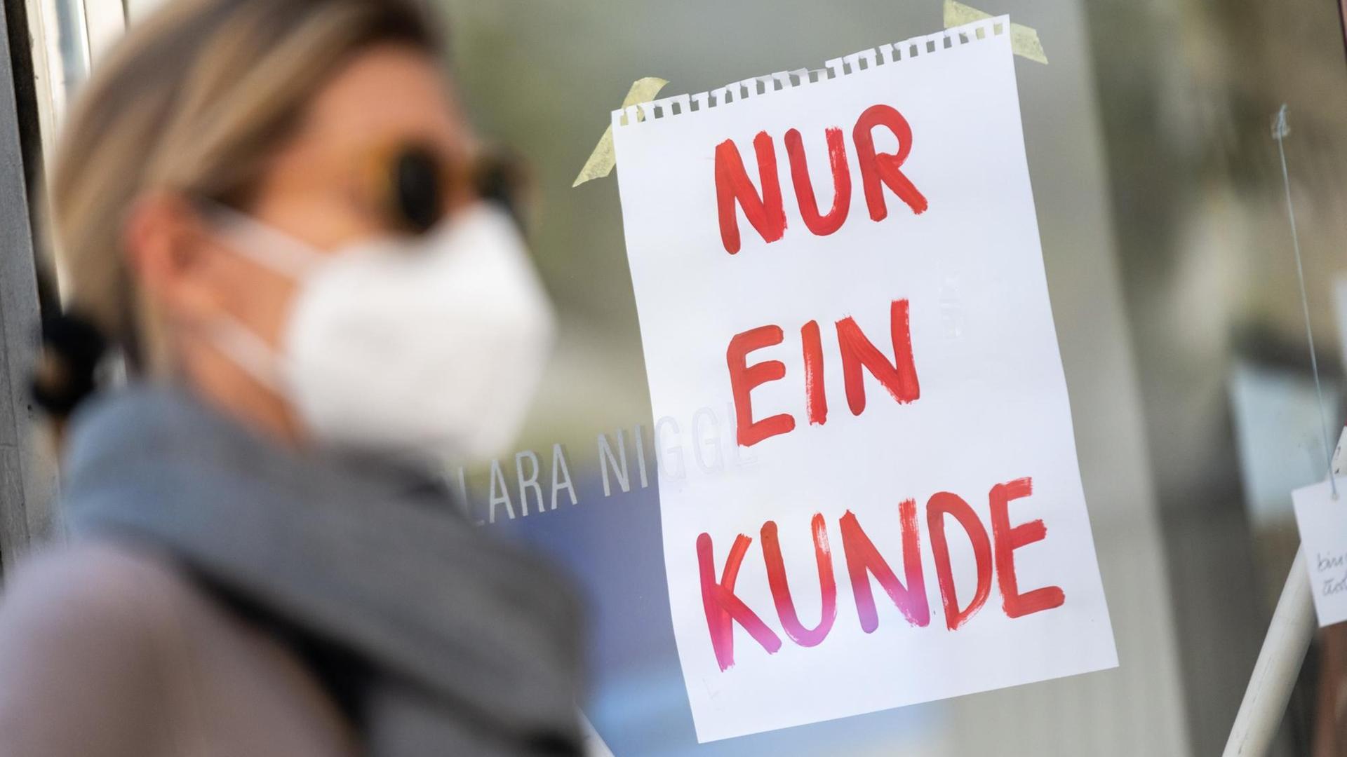 Eine Frau mit Nase-Mund-Schutzmaske geht in München an einem Geschäft vorbei, an dem ein Schild mit der Aufschrift "Nur ein Kunde" angebracht ist. Eine Frau mit Nase-Mund-Schutzmaske geht in München an einem Geschäft vorbei, an dem ein Schild mit der Aufschrift "Nur ein Kunde" angebracht ist.