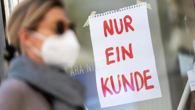 Eine Frau mit Nase-Mund-Schutzmaske geht in München an einem Geschäft vorbei, an dem ein Schild mit der Aufschrift "Nur ein Kunde" angebracht ist.