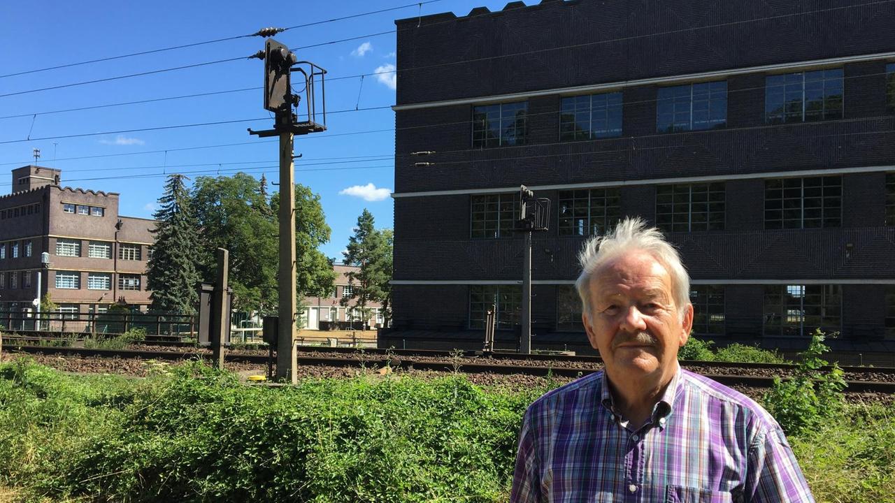 Ein älterer Mann in einem karierten Kurzarmhemd steht vor einem Bahngleis. Im Hintergrund dunkel geklinkerte Industriebauten. Ein älterer Mann in einem karierten Kurzarmhemd steht vor einem Bahngleis. Im Hintergrund dunkel geklinkerte Industriebauten.