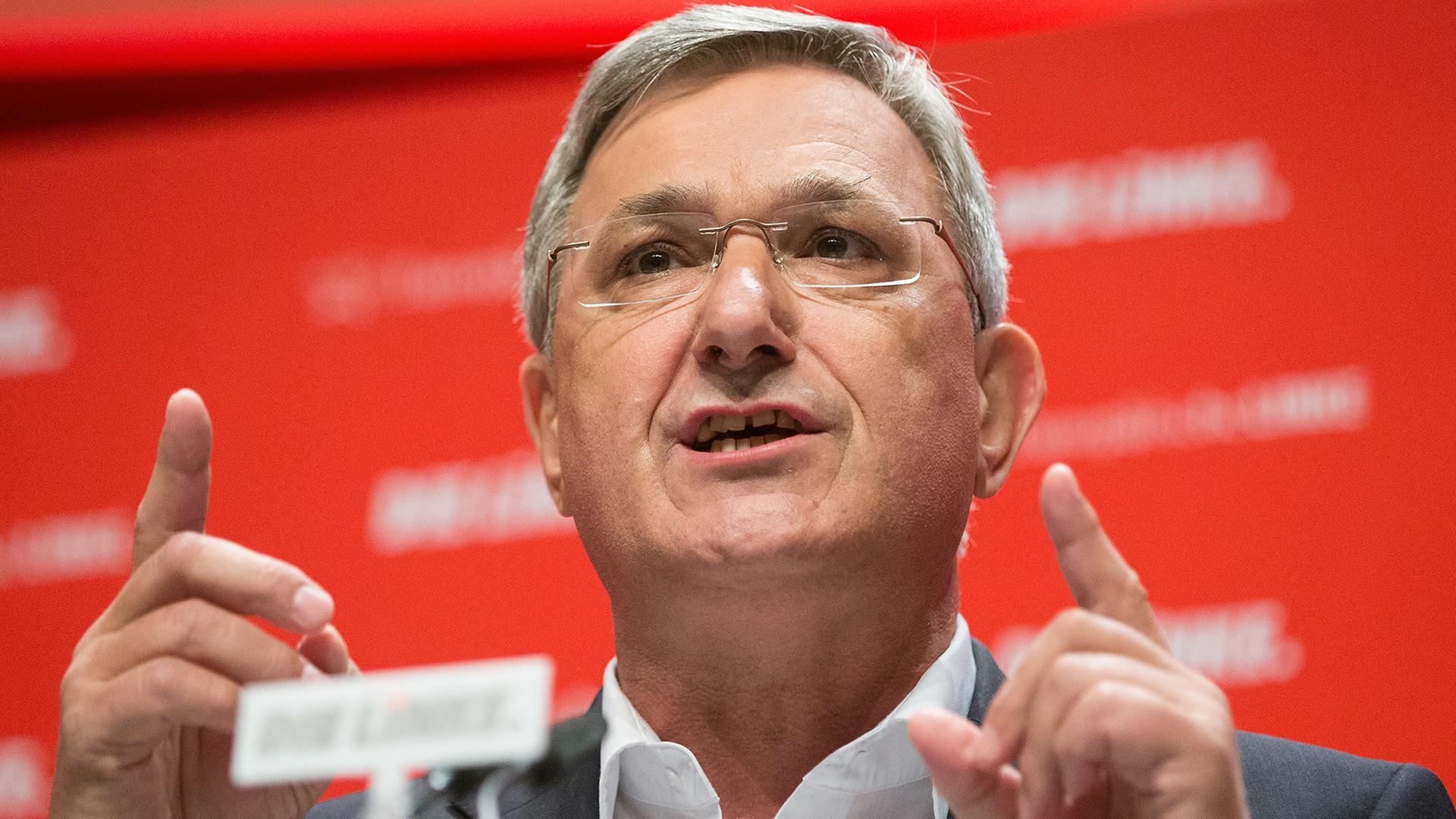 Der Vorsitzende der Partei Die Linke, Bernd Riexinger, steht vor dem Parteilogo am Rednerpult und hebt beide Zeigefinger.