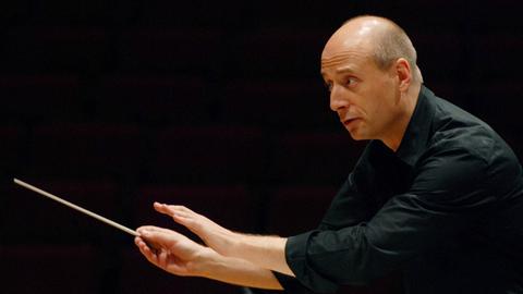 Ein Dirigent fordert mit dämpfender Geste Orchestermusiker zu leisem Spielen auf.