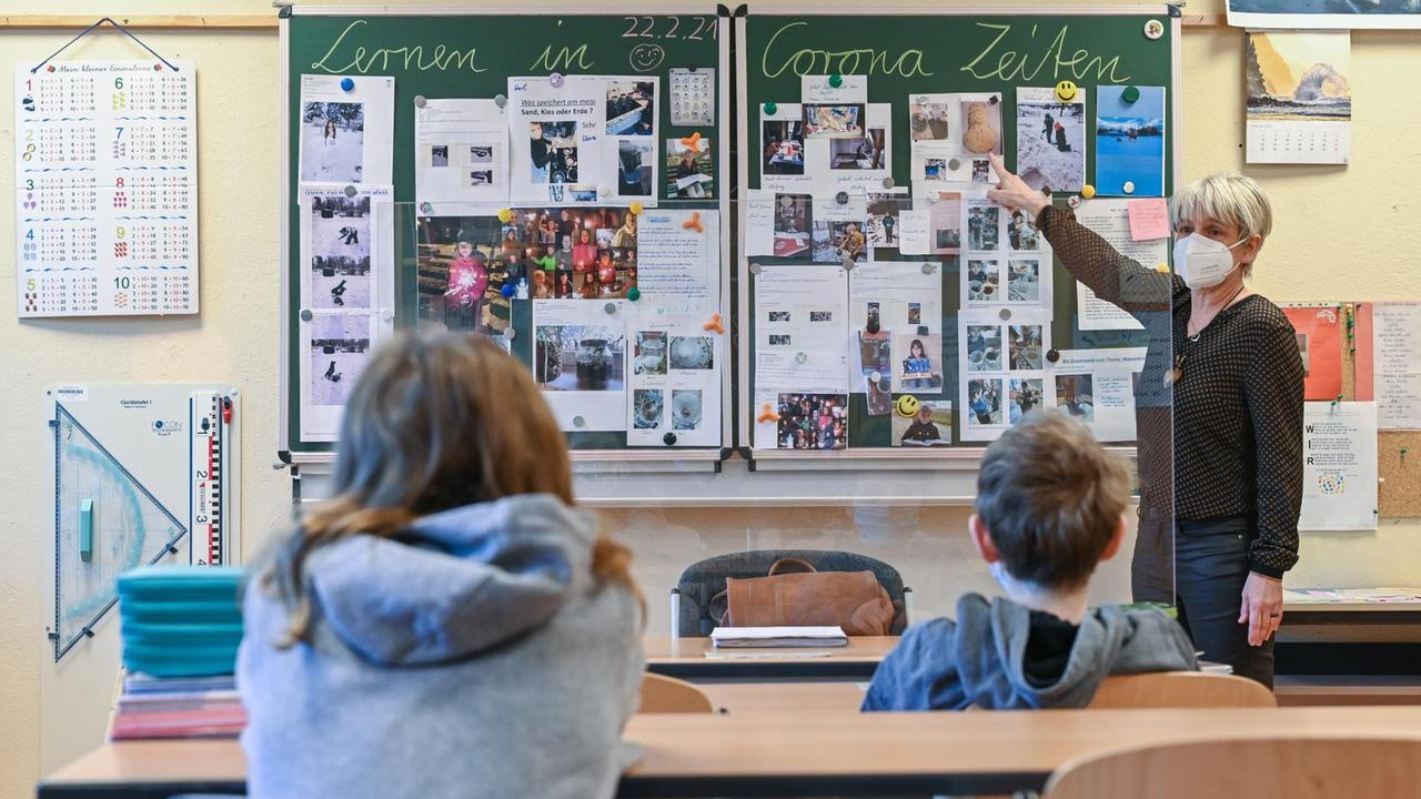 Unterrichtsszene: In einem Klassenzimmer sieht man zwei Kinder von hinten. Sie sitzen jeweils einzeln an einer Bank und blicken nach vorne zur Tafel, neben der ihre Lehrerin steht. Unterrichtsszene: In einem Klassenzimmer sieht man zwei Kinder von hinten. Sie sitzen jeweils einzeln an einer Bank und blicken nach vorne zur Tafel, neben der ihre Lehrerin steht.