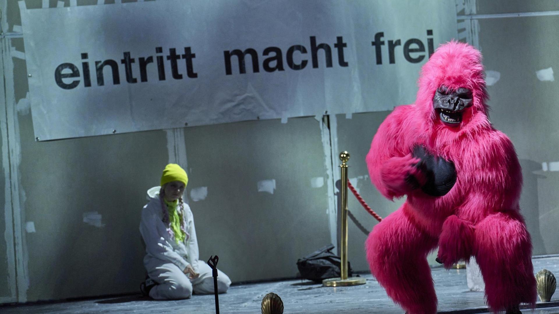 Szenenfoto: Ein Mensch verkleidet als pinkfarbener Gorilla blickt Rochtung Bühne. Im Hintergrund ein großes Plakat mit der Aufschrift: "Eintritt macht frei". Unter dem Plakat sitzt eine Jugendliche, die wohl Greta Thunberg darstellen soll. Szenenfoto: Ein Mensch verkleidet als pinkfarbener Gorilla blickt Rochtung Bühne. Im Hintergrund ein großes Plakat mit der Aufschrift: "Eintritt macht frei". Unter dem Plakat sitzt eine Jugendliche, die wohl Greta Thunberg darstellen soll.