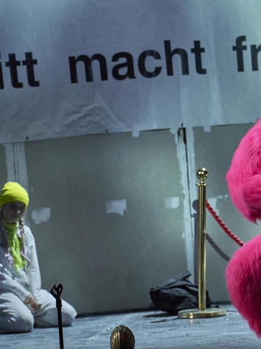 Szenenfoto: Ein Mensch verkleidet als pinkfarbener Gorilla blickt Rochtung Bühne. Im Hintergrund ein großes Plakat mit der Aufschrift: "Eintritt macht frei". Unter dem Plakat sitzt eine Jugendliche, die wohl Greta Thunberg darstellen soll.