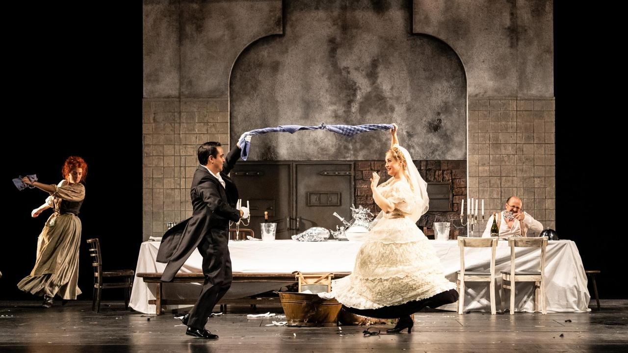 In einer Küchenkulisse auf der Bühne tanzt im Vordergrund ein Hochzeitspaar, im Hintergrund tanzt eine rothaarige Frau und ein älterer Man isst an einer Tafel sitzend. In einer Küchenkulisse auf der Bühne tanzt im Vordergrund ein Hochzeitspaar, im Hintergrund tanzt eine rothaarige Frau und ein älterer Man isst an einer Tafel sitzend.