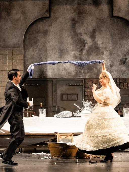 In einer Küchenkulisse auf der Bühne tanzt im Vordergrund ein Hochzeitspaar, im Hintergrund tanzt eine rothaarige Frau und ein älterer Man isst an einer Tafel sitzend.
