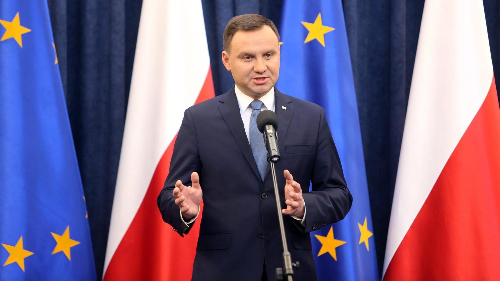Andrzej Duda spricht bei einer Pressekonferenz in ein Mikrofon, im Hintergrund die Flaggen Polens und der EU.