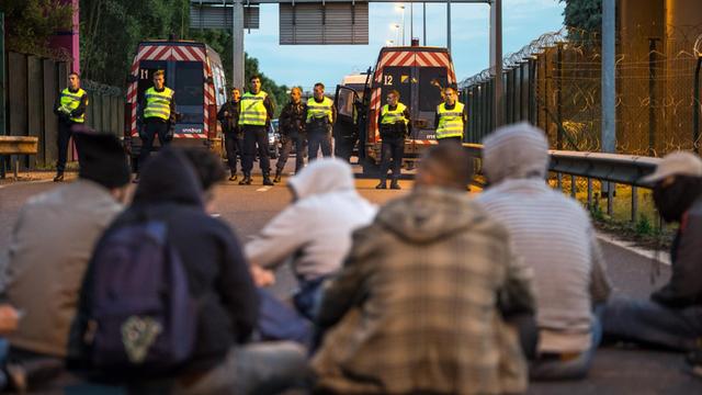 Flüchtlinge sitzen am Eurotunnel in Calais. Der Weg wird von Polizisten blockiert.