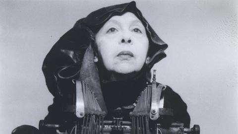 Das Kunstwerk "Mrs Oliver in her traveling costume" von Geta Brătescu. Ein Schwarz-Weiss-Foto das die in ganz in schwarz gekleidete Künstlerin mit einer mechanischen Schreibmaschine zeigt.