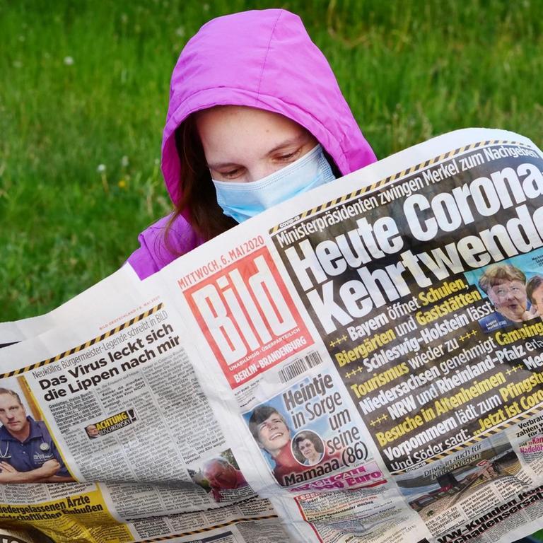 Eine Frau trägt eine Maske zum Schutz gegen das neuartige Coronavirus, während sie auf einer Bank sitzt und die Bildzeitung liest.Die Titelschlagzeile lautet: "Heute Corona-Kehrtwende!".