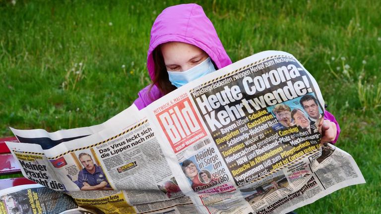 Eine Frau trägt eine Maske zum Schutz gegen das neuartige Coronavirus, während sie auf einer Bank sitzt und die Bildzeitung liest.Die Titelschlagzeile lautet: "Heute Corona-Kehrtwende!".