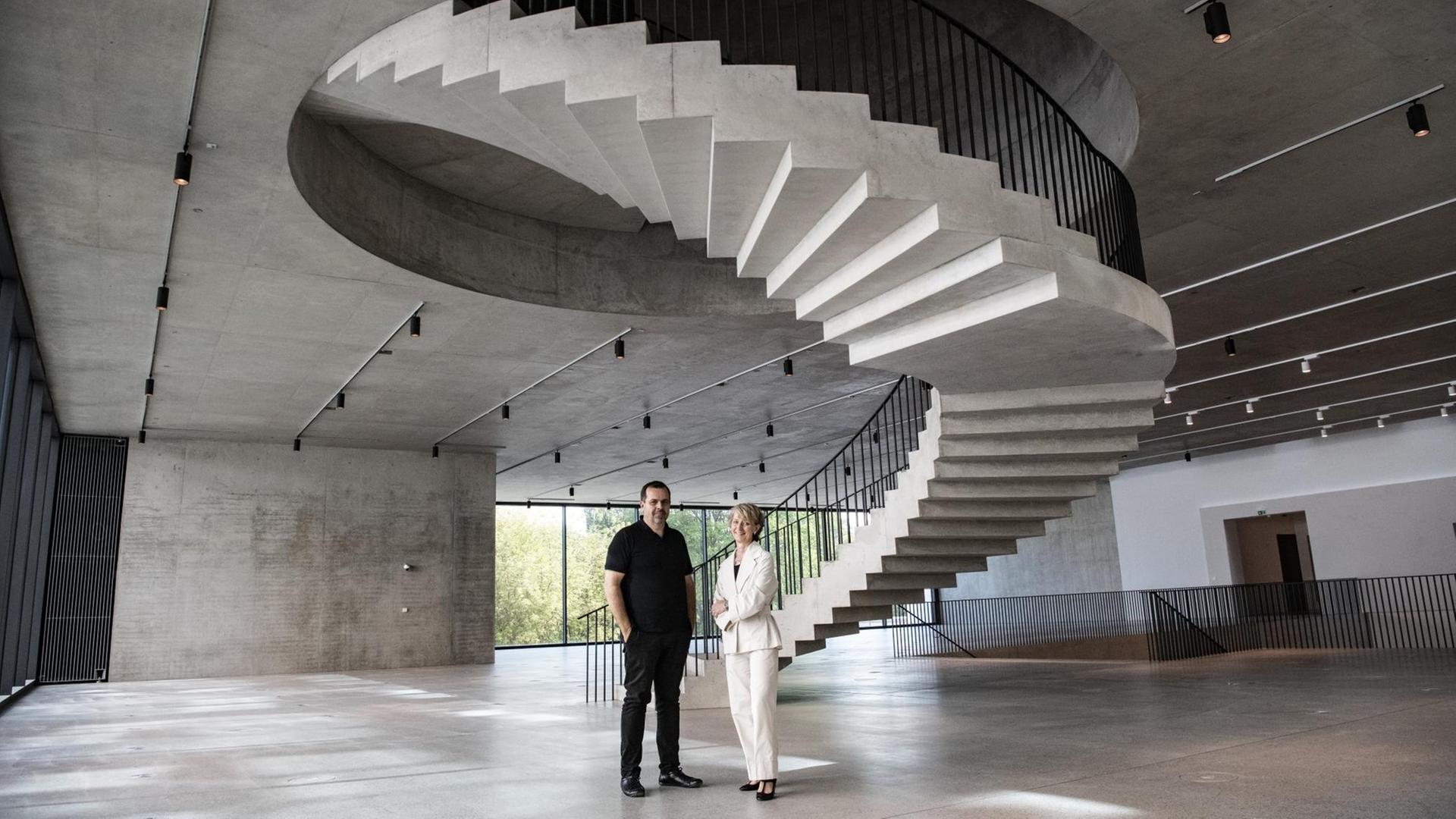 Die beiden stehen in einem Obergeschoß aus Beton, in dessen Mitte eine spektakuläre Wendeltreppe in den Stock darüber ragt. Die beiden stehen in einem Obergeschoß aus Beton, in dessen Mitte eine spektakuläre Wendeltreppe in den Stock darüber ragt.