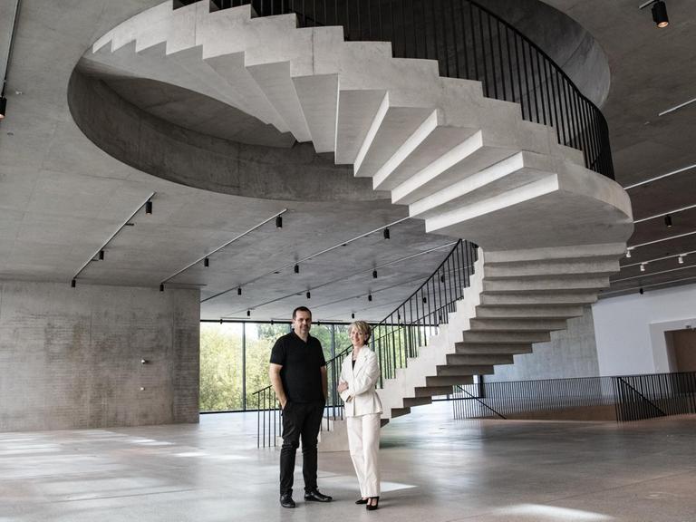 Die beiden stehen in einem Obergeschoß aus Beton, in dessen Mitte eine spektakuläre Wendeltreppe in den Stock darüber ragt.