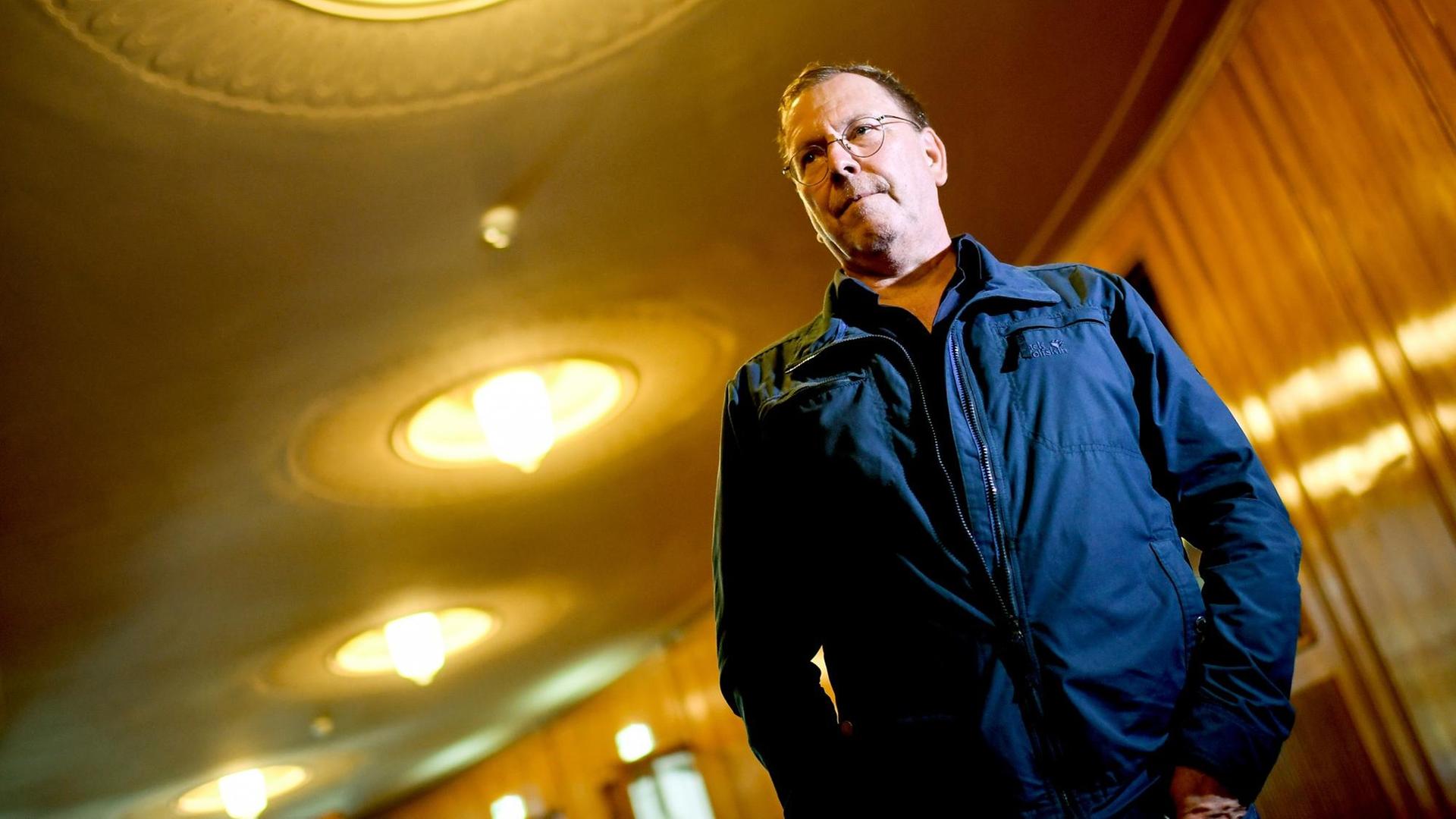 René Pollesch steht in einem Gang mit braunen Holzwänden und runden Leuchtern an der Decke. Er ist von schräg unten fotografiert und schaut an der Kamera vorbei. René Pollesch steht in einem Gang mit braunen Holzwänden und runden Leuchtern an der Decke. Er ist von schräg unten fotografiert und schaut an der Kamera vorbei.