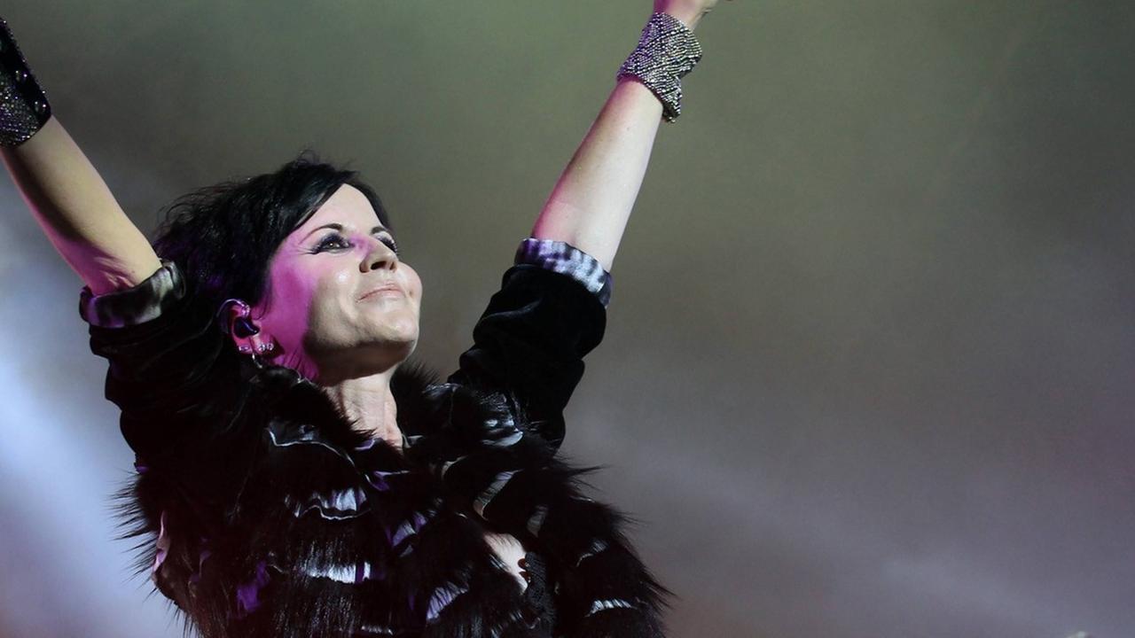 Dolores O’Riordan steht mit nach oben gereckten Armen auf der Bühne, in einer Hand hält sie ein Mikrofon. Dolores O’Riordan steht mit nach oben gereckten Armen auf der Bühne, in einer Hand hält sie ein Mikrofon.