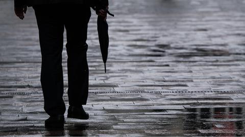 Die Beine eines Passanten auf einer regennassen Straße sind zu sehen. Er trägt einen Regenschirm.