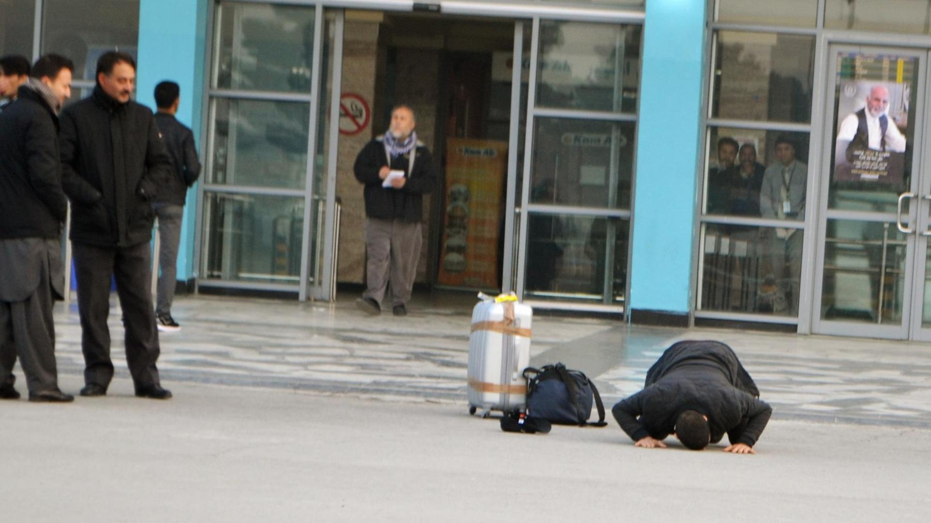 Ein Mann sitzt gebeugt auf der Straße und küsst den Boden, neben ihm ein silberner Koffer. Im Hintergrund das Flughafengebäude.