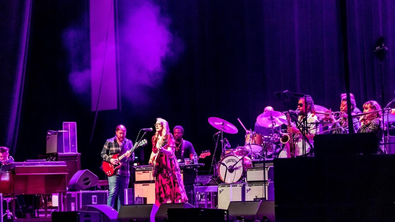Auf einer Bühne: Eine blonde Frau in rotem Kleid steht an einem Mikrofon und singt hinein während sie Gitarre spielt. Neben und hinter ihr weitere Musiker.