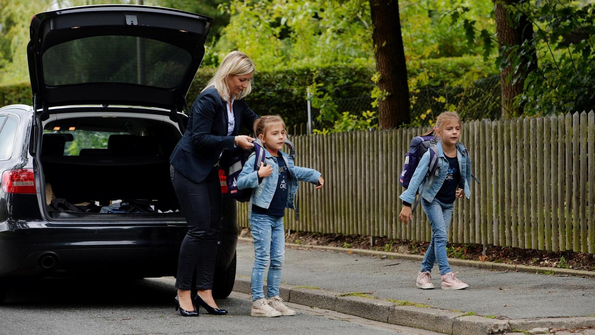 Eine Mutter in Businesskleidung hilft ihren Töchtern vor dem Auto, die Schulranzen aufzuziehen Eine Mutter in Businesskleidung hilft ihren Töchtern vor dem Auto, die Schulranzen aufzuziehen