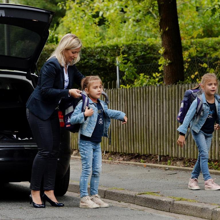 Eine Mutter in Businesskleidung hilft ihren Töchtern vor dem Auto, die Schulranzen aufzuziehen 