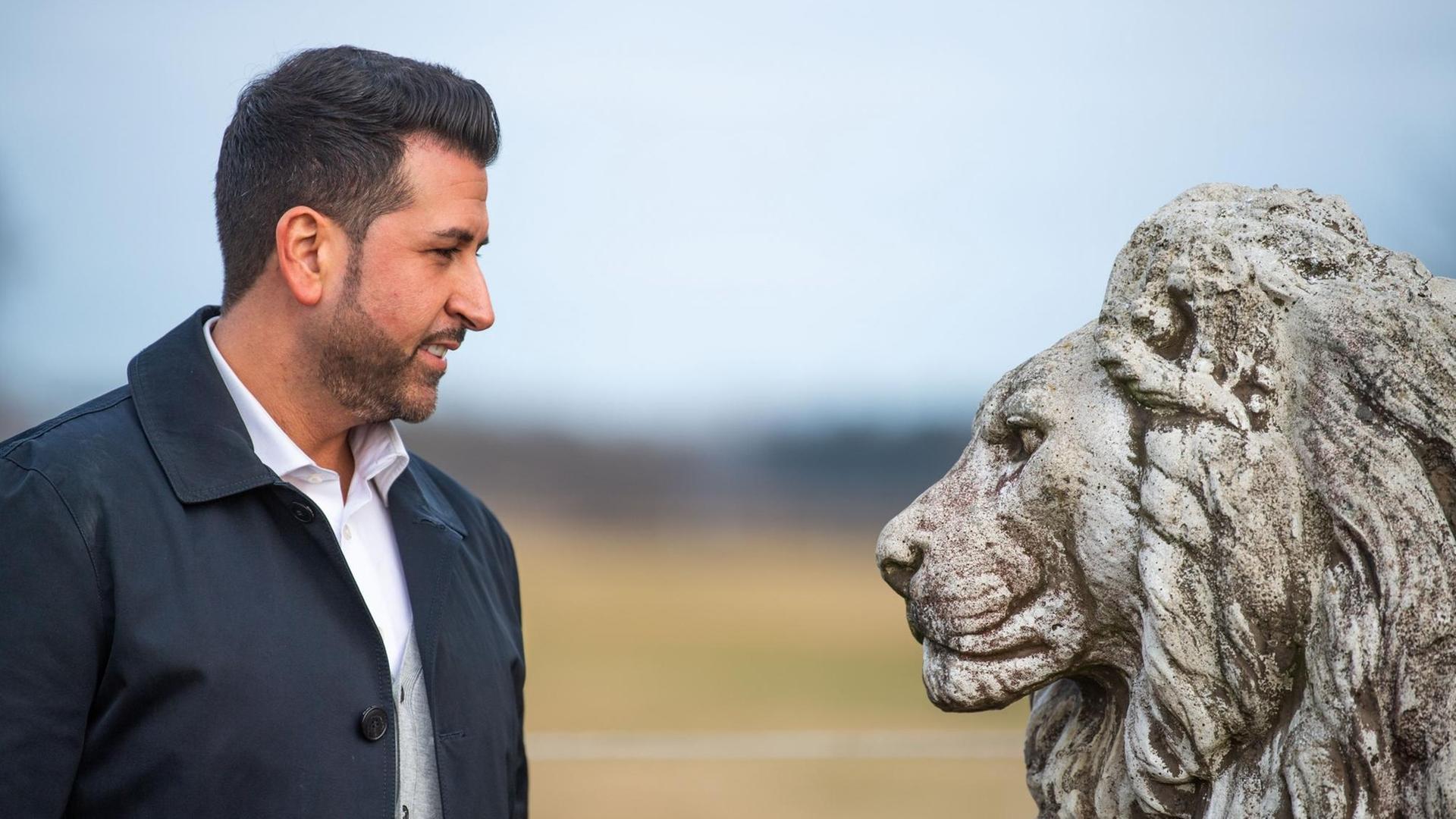Ozan Iyibas steht einem Löwenkopf aus Stein gegenüber. Ozan Iyibas steht einem Löwenkopf aus Stein gegenüber.