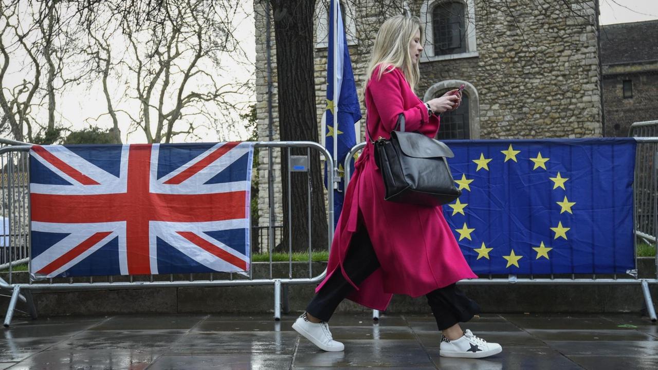 Eine Frau in einem roten Mantel geht in London an einer Absperrung vorbei, an der eine britische und eine EU-Flagge hängen. Eine Frau in einem roten Mantel geht in London an einer Absperrung vorbei, an der eine britische und eine EU-Flagge hängen.
