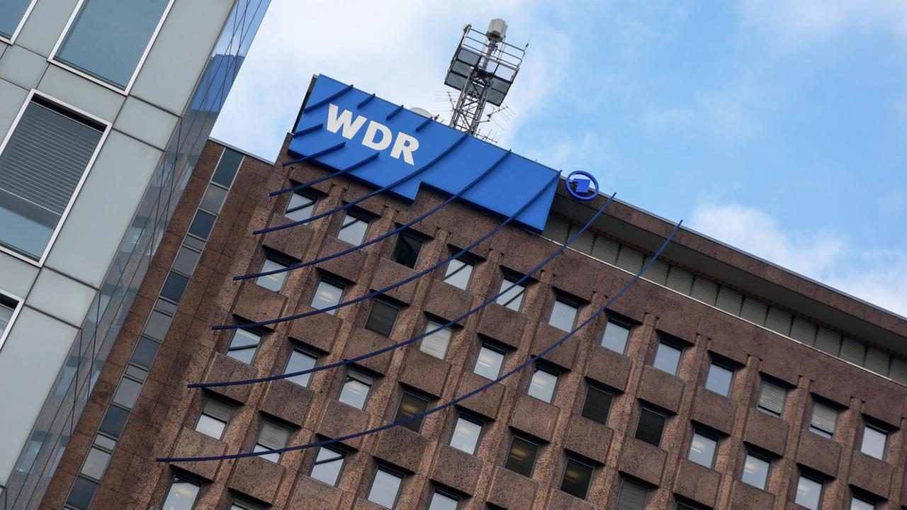 Das Bild zeigt einen Teil des WDR-Komplexes. An der oberen linken Ecke sieht man den blauen Schriftzug des Senders mit blauen Strichen, die - Sendewellen darstellend - im Halbkreis über mehrere der Fenster verlaufen.