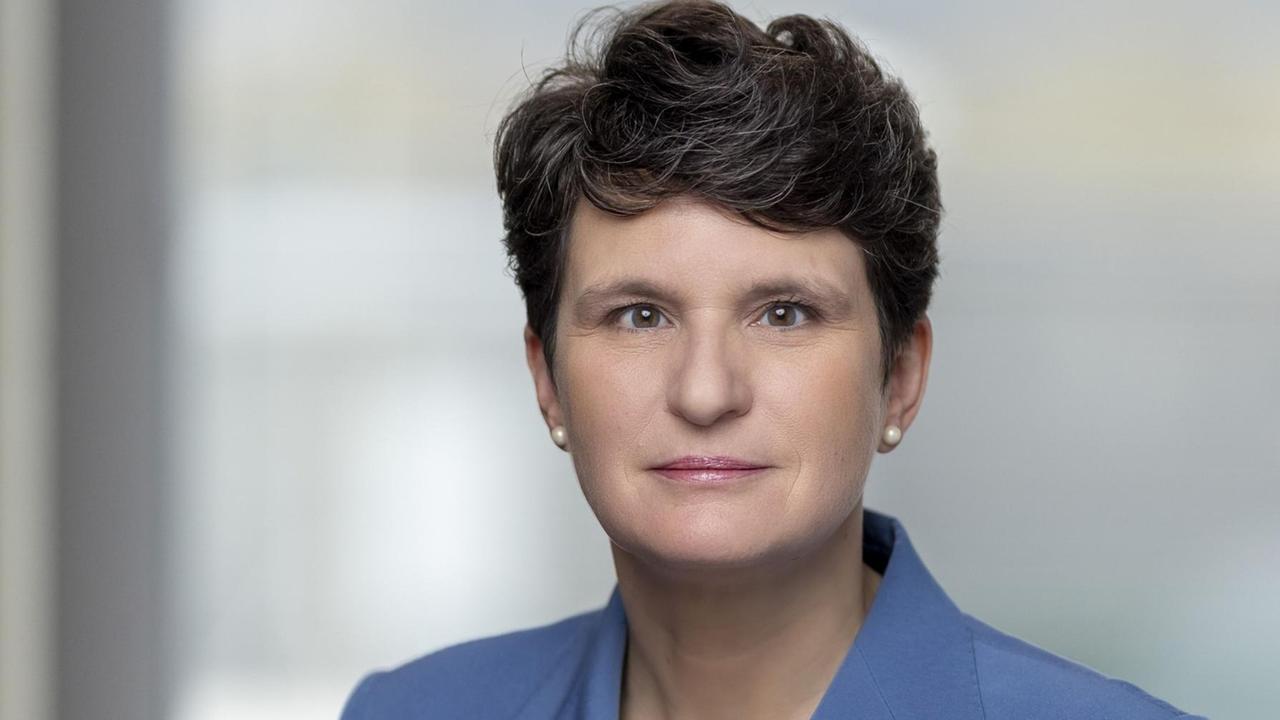 Eine Frau mit kurzen, lockigen, dunklen Haaren schaut in die Kamera. Sie trägt einen blauen Blazer. Eine Frau mit kurzen, lockigen, dunklen Haaren schaut in die Kamera. Sie trägt einen blauen Blazer.