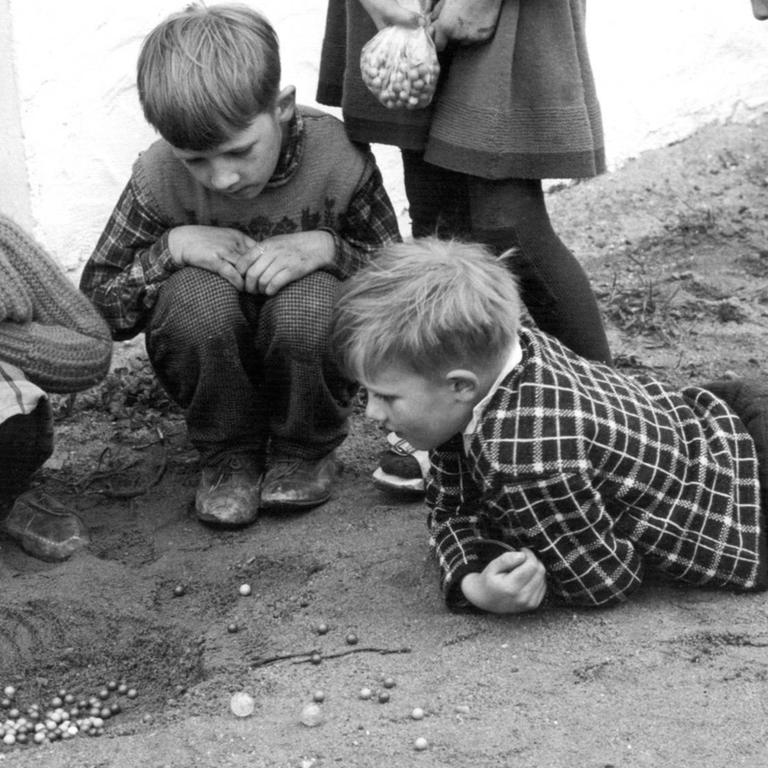 Fünf Kinder spielen im Sand mit Murmeln.