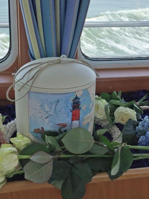 Urne und Blumen vor einem Schiffsfenster mit Blick auf das Meer.