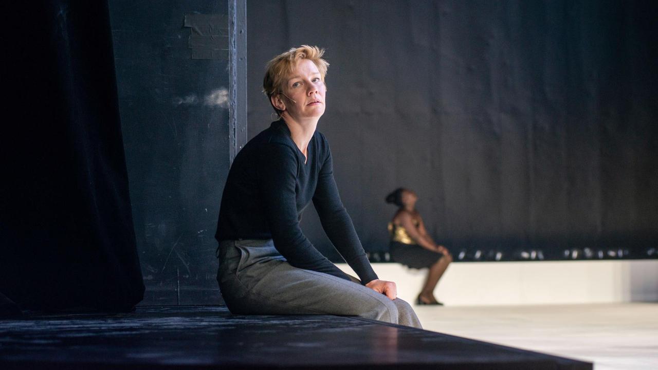 Sandra Hüller in der Rolle von Hamlet sitzt auf einem Podest und blickt seitlich in die Kamera. Im Hintergrund ist Mercy Dorcas Otieno zu sehen. Sandra Hüller in der Rolle von Hamlet sitzt auf einem Podest und blickt seitlich in die Kamera. Im Hintergrund ist Mercy Dorcas Otieno zu sehen.