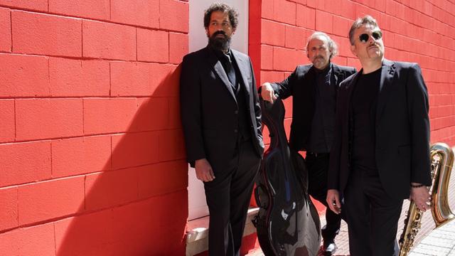 Das Trio Camerata Flamenco Project posiert in schwarzen Anzügen vor einer roten Wand