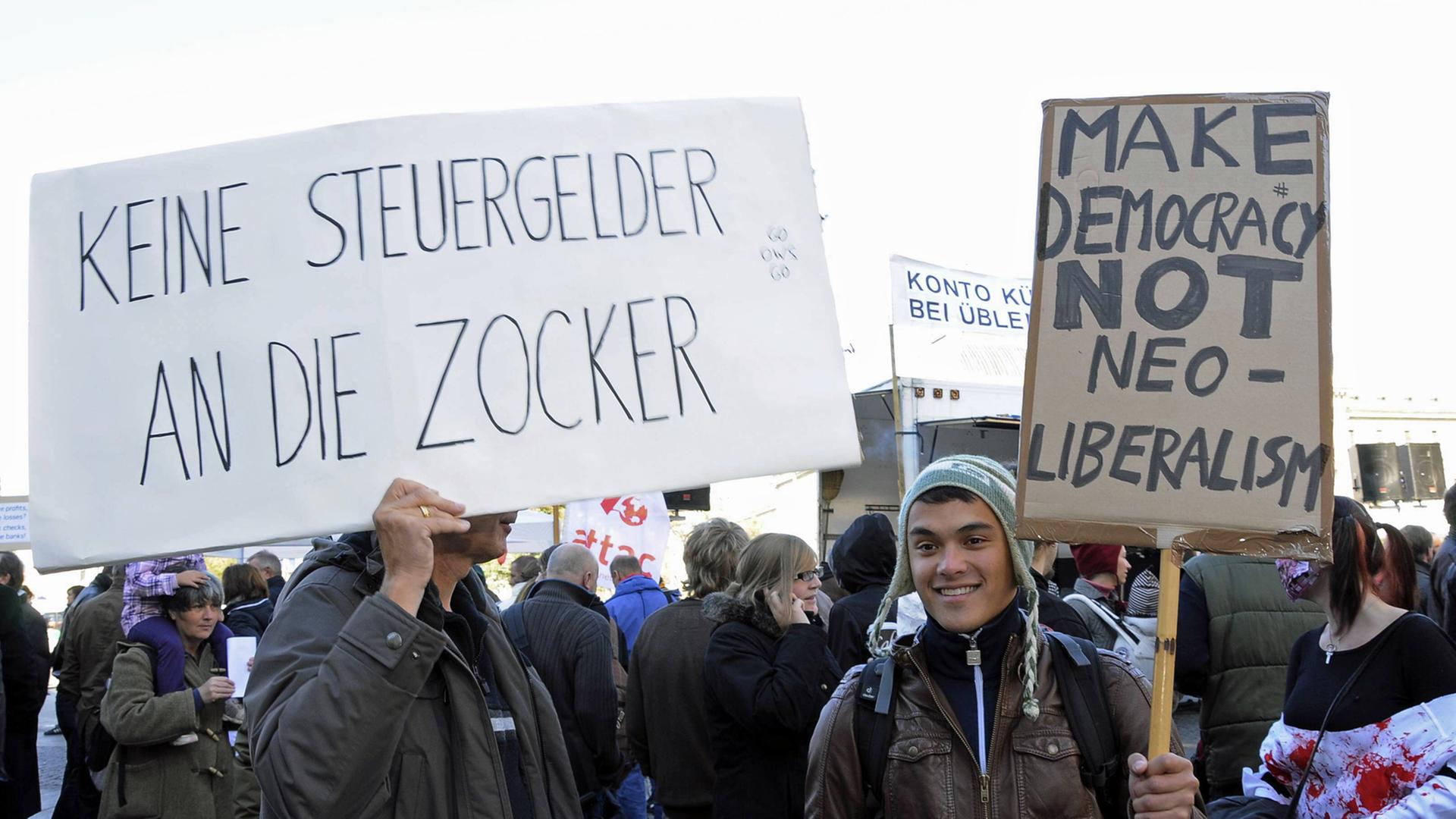 "Make Democracy not Neoliberalism" und "Keine Steuergelder an die Zocker" steht auf zwei Plakaten bei einer Demonstration gegen die Ungerechtigkeit des Finanzsystems und für mehr Demokratie in München im Oktober 2011
