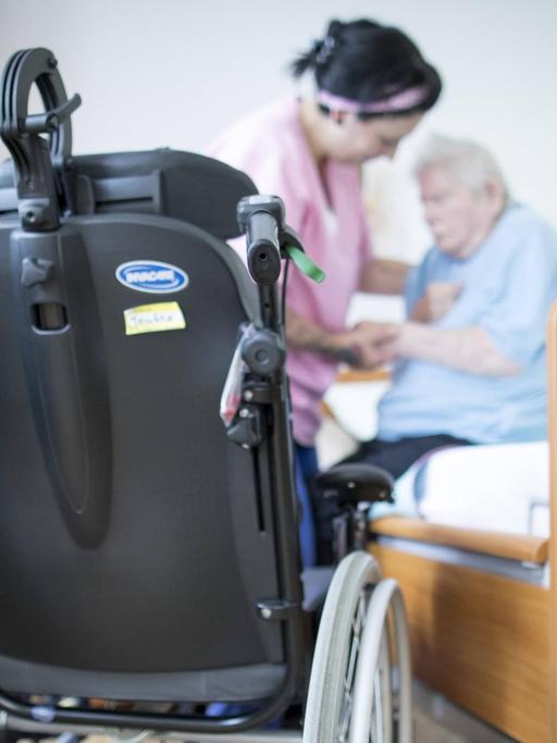 Eine Altenpflegerin hilft einer Bewohnerin in einem Altenpflegeheim aus dem Bett. Im Vordergrund steht ein Rollstuhl.