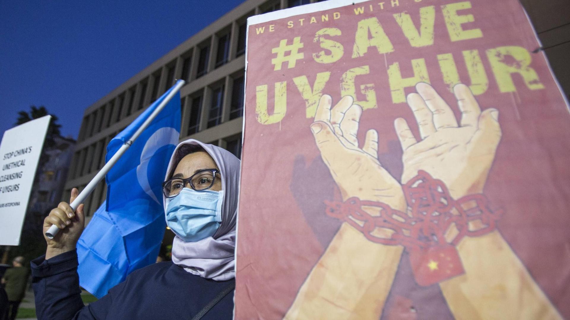 Eine Frau mit blauer Fahne steht neben einem Plakat, auf dem "#Save Uyghur" steht.