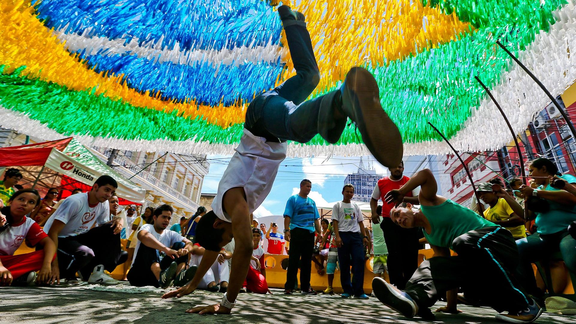 Unter einer brasilianischen Flagge tanzen zwei junge Männer Capoeira: Der eine steht gerade auf seinen Händen, der andere dreht sich am Boden um sich selbst. Unter einer brasilianischen Flagge tanzen zwei junge Männer Capoeira: Der eine steht gerade auf seinen Händen, der andere dreht sich am Boden um sich selbst.